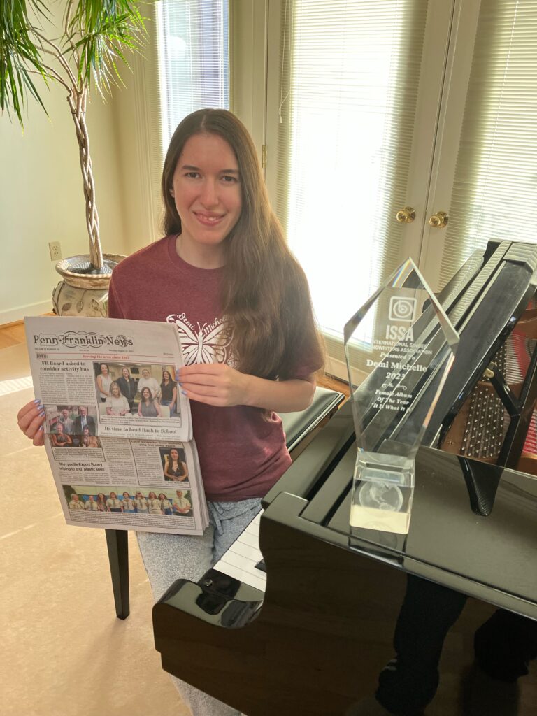 Demi Michelle is sitting on her piano bench holding the Penn-Franklin News, featuring her on the front cover. Her ISSA Awards crystal is on the piano.