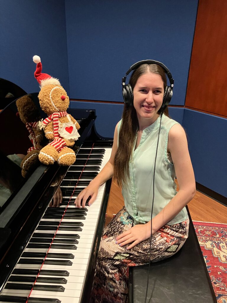 Demi Michelle is sitting at the piano, smiling and wearing headphones. Her gingerbread is on the piano, bringing the festive magic.
