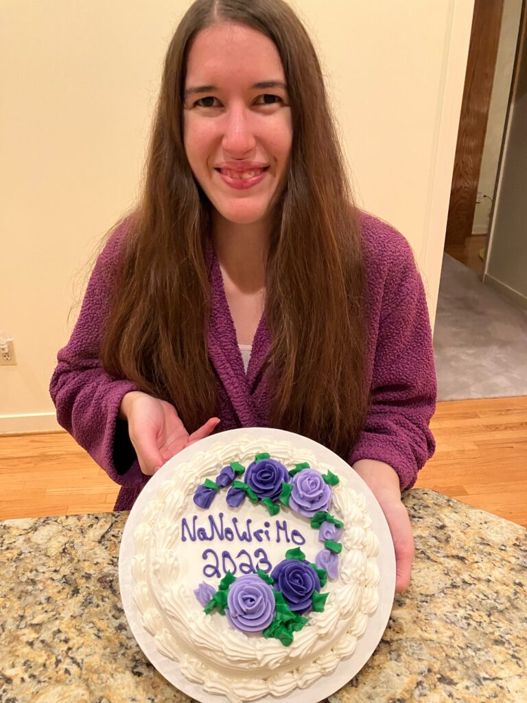Demi is smiling with her cake, which says NaNoWriMo 2023 in purple icing with flowers