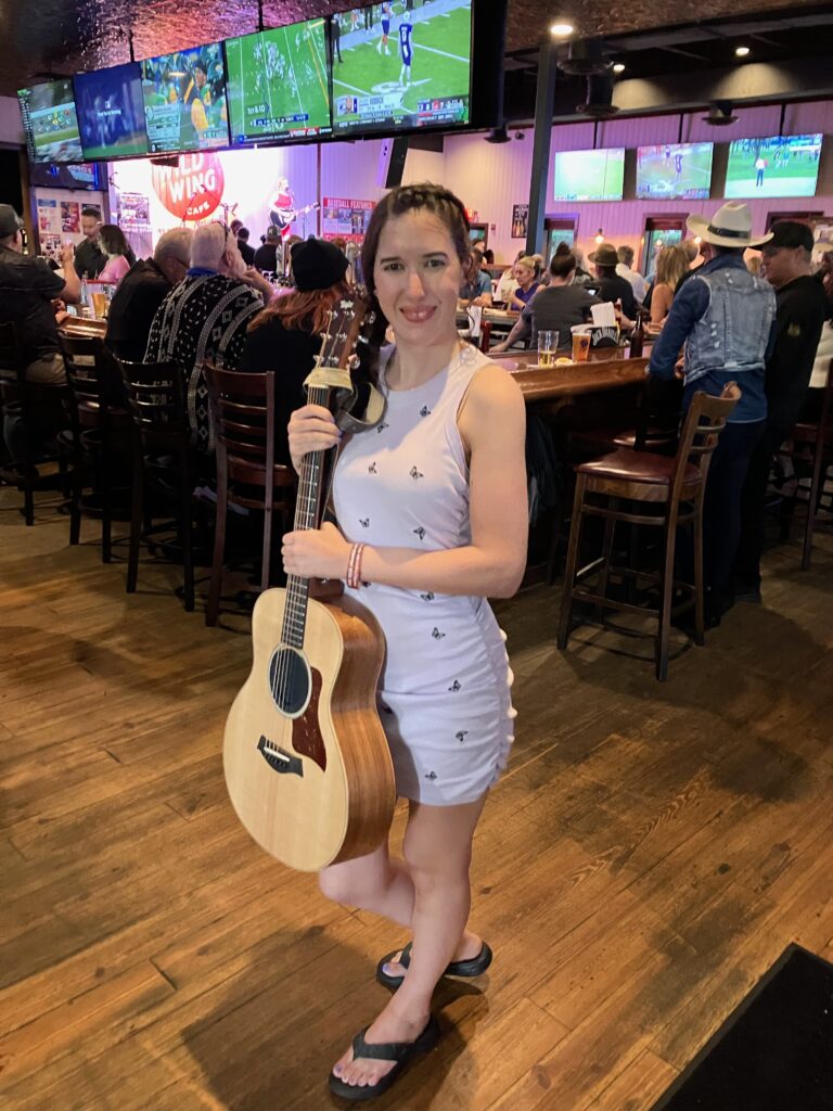 Demi Michelle smiling with her guitar in Wild Wing Cafe and wearing a butterfly dress