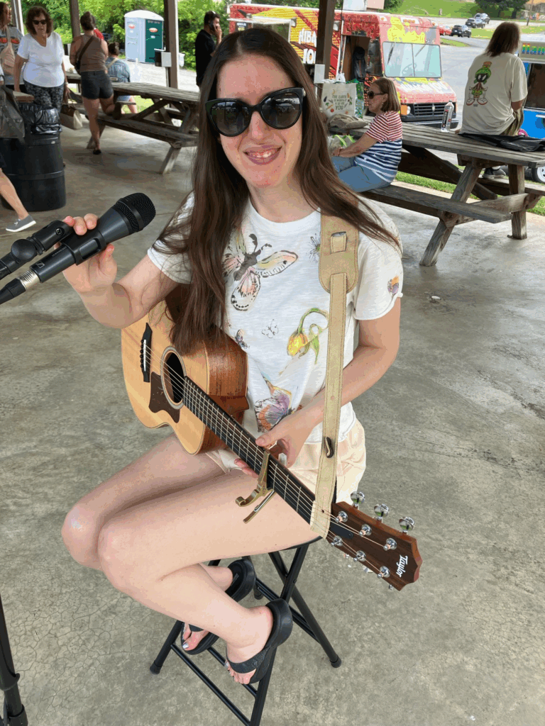 Demi smiling by her microphone with her guitar at the farmers market